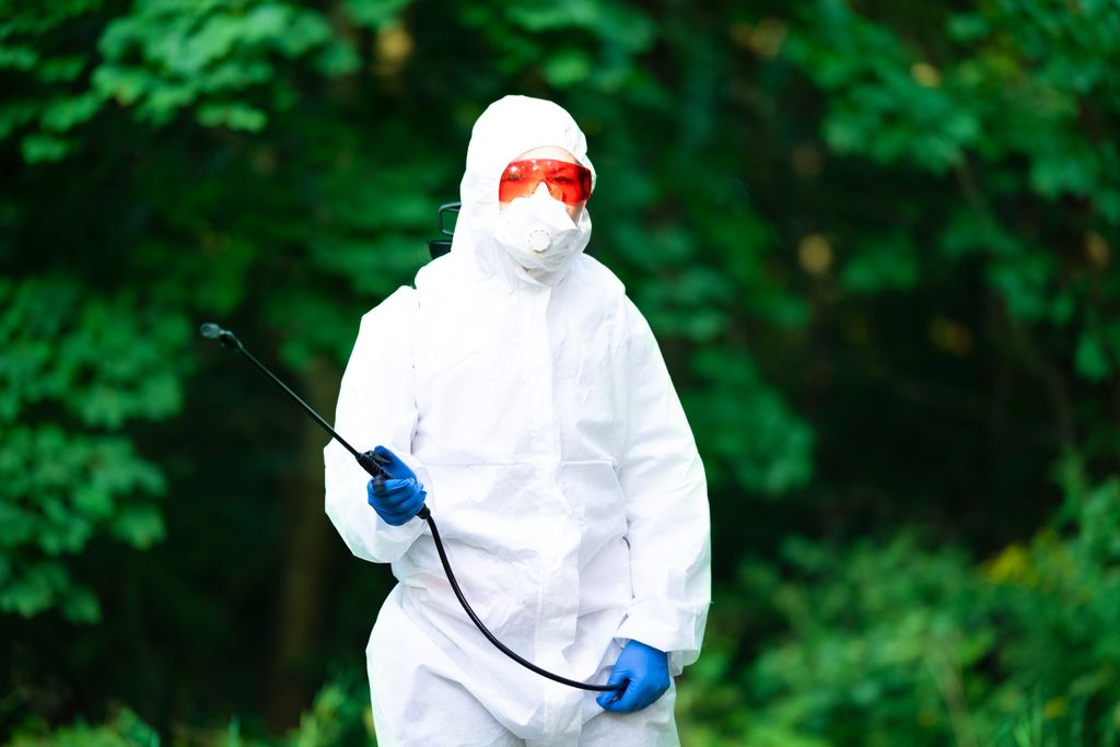 An operator in a hooded white protective coverall, safety goggles, respirator mask and nitrile gloves, holding a sprayer lance in front of green foliage