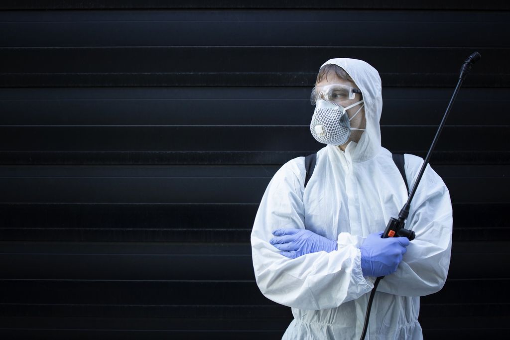 An operator in a hooded white protective coverall, safety goggles, respirator mask and nitrile gloves, arms crossed, holding the lance of a knapsack sprayer