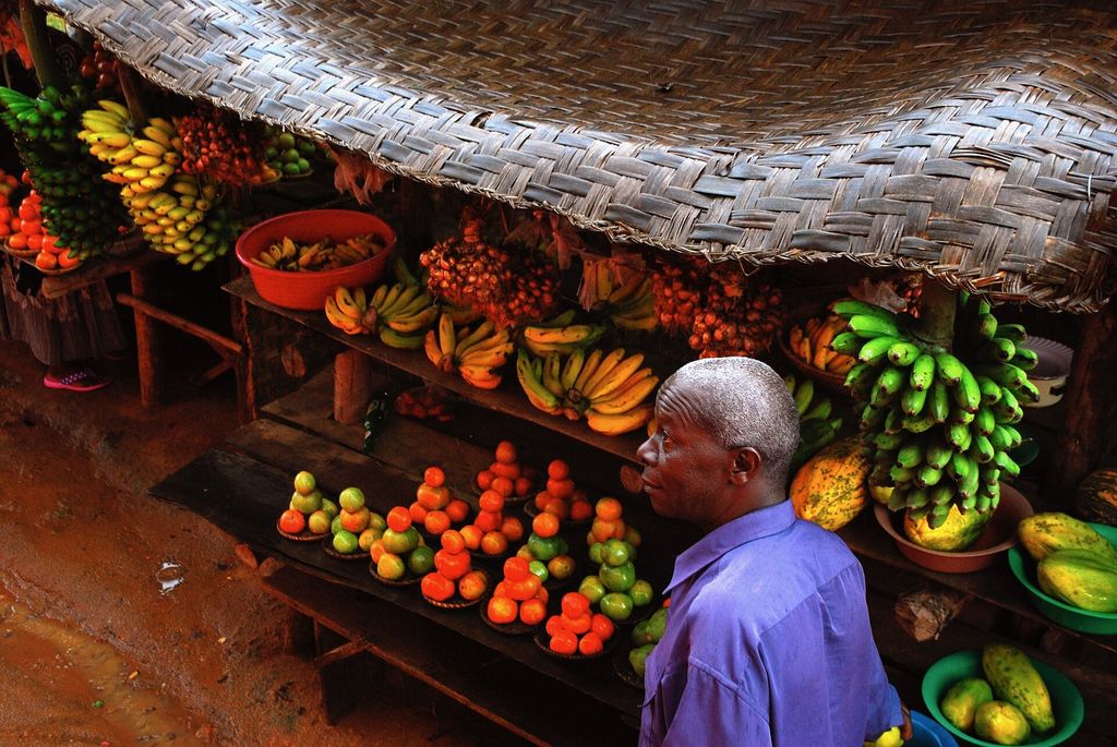 A roadside stall under a woven roof piled with bananas, matooke, mangoes, oranges and tomatoes, with a vendor in front